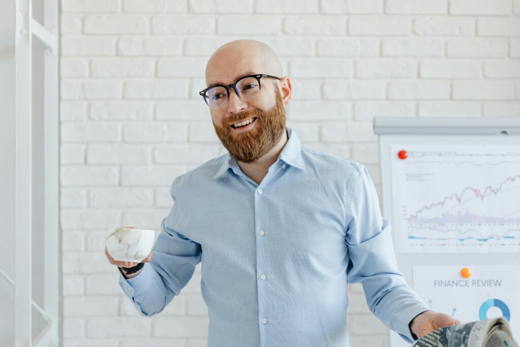 pexels-photo-7876976-7876976 Smiling businessman holding coffee cup and newspaper in office with chart in background.